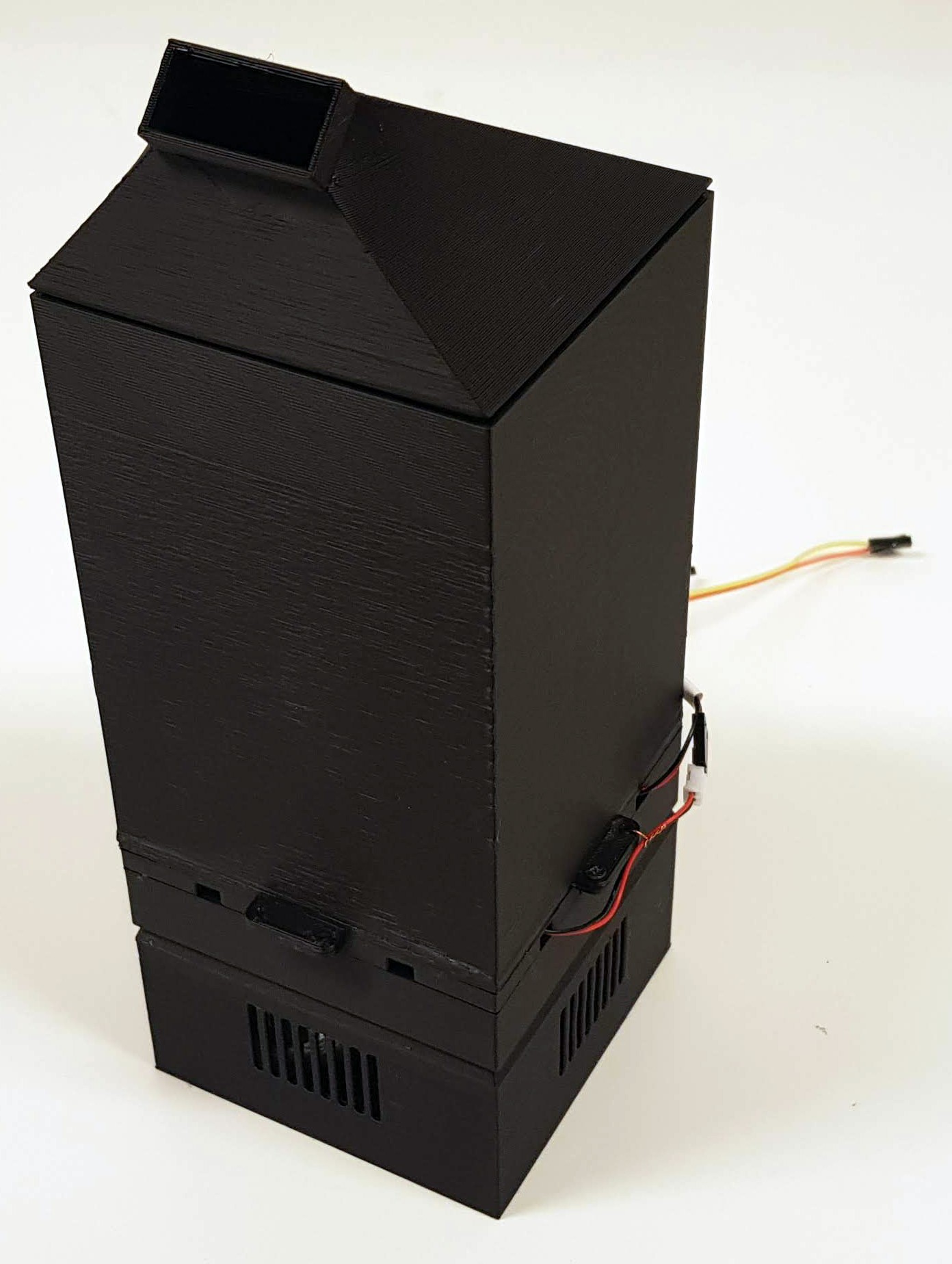 The Scentony device fully assembled, standing. It's a black 3d printed box with groves at the bottom and a slightly tilted funnel attachment at the top